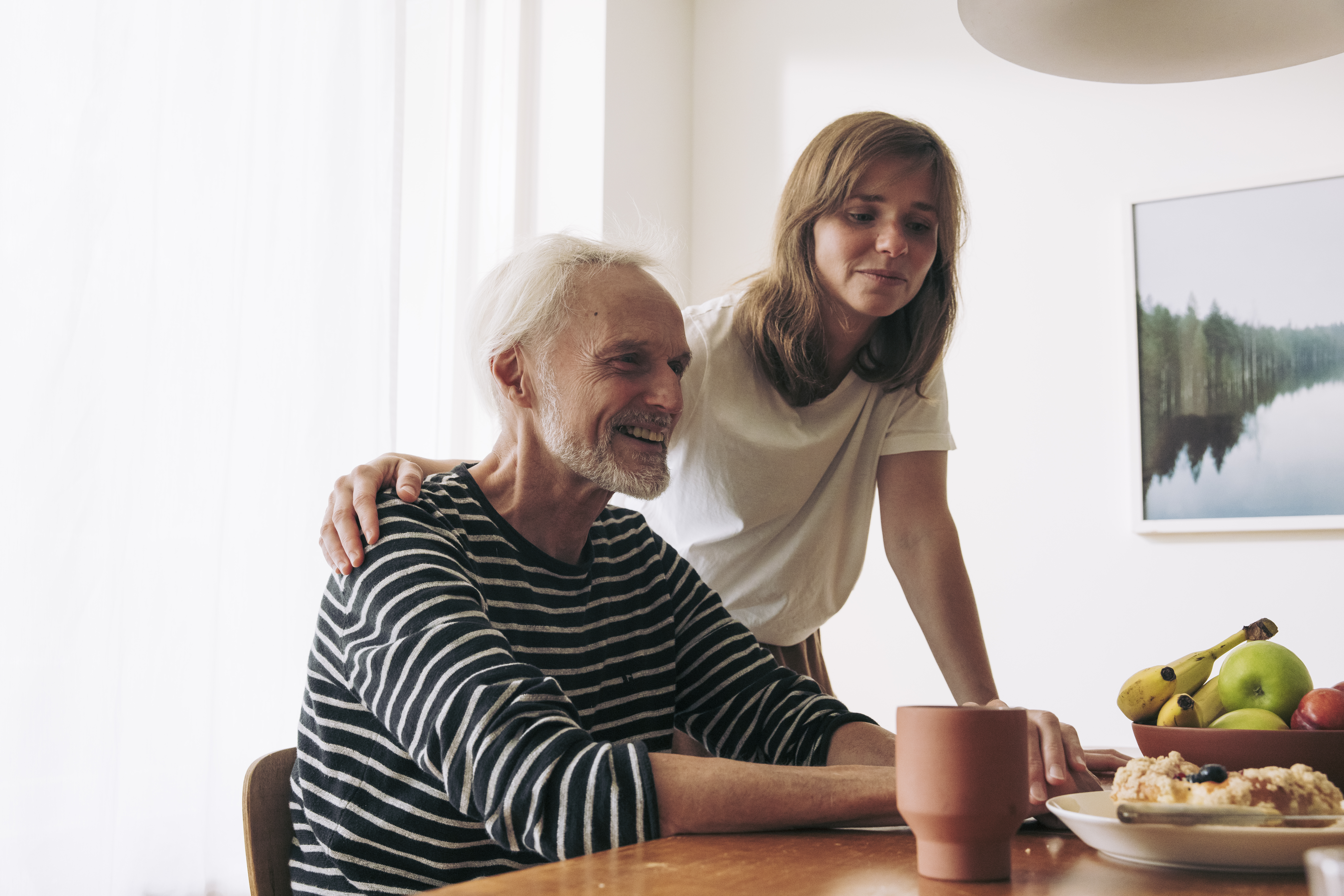 Kuvassa vanhempi mies istuu pöydän ääressä ja nuorempi nainen seisoo hänen vierellään. Hänen kätensä lepää miehen olkapäällä.